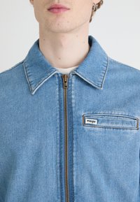 Jasnoniebieska kurtka dżinsowa z kołnierzykiem, zamkiem błyskawicznym z przodu i widocznym szwem. Posiada małą naszywkę z napisem "Wrangler" na kieszeni na piersi.