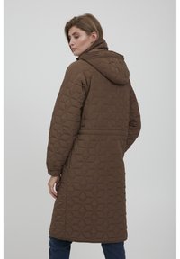 Kvinde iført en lang brun vatteret frakke med hætte, der står lidt drejet til siden mod en ensfarvet lys baggrund.