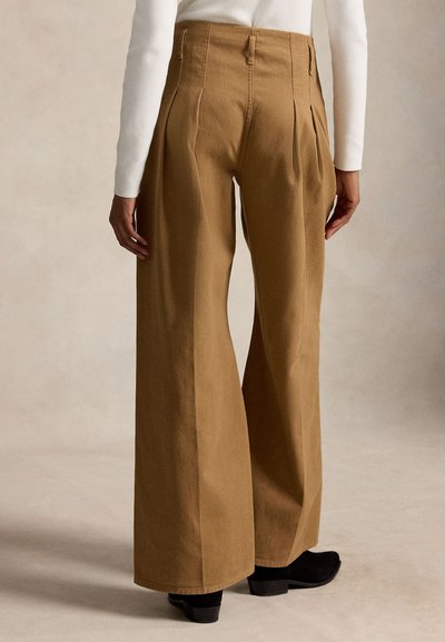 Vue de dos d'une personne portant un pantalon marron taille haute et à jambes larges, un haut blanc à manches longues et des chaussures à talons noires sur fond neutre.
