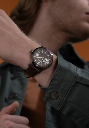 Montre avec bracelet en cuir marron, cadran analogique noir, accents argentés et marqueurs d'heure. Elle dispose d'une fenêtre pour la date et d'un boîtier circulaire.