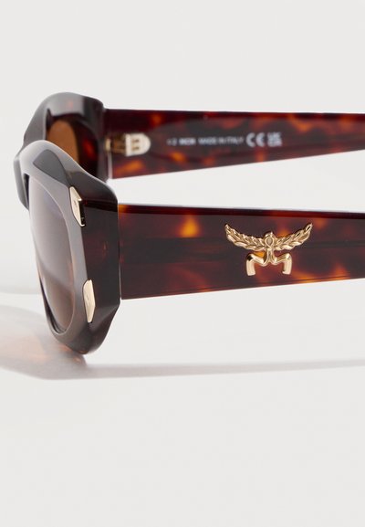 Γυαλιά ηλίου με σκελετό χελώνας, παχιά τετράγωνα πλαίσια και χρυσό έμβλημα με φτερά στο πλευρικό βραχίονα.