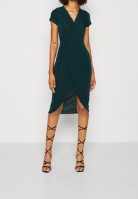Donna che indossa un vestito avvolgente di lunghezza al ginocchio di un verde scuro, con maniche corte e sandali con tacco alto, alla caviglia, neri con lacci, in piedi su uno sfondo bianco.