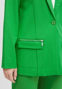 Blazer verde hecho de tela texturizada con un solo botón verde y un detalle de bolsillo con cremallera en la parte frontal, combinado con pantalones verdes a juego.