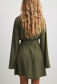 Frau mit langen, lockigen blonden Haaren, die ein mit einem Gürtel versehenes olivgrünes langärmliges Kleid trägt, von hinten vor einem schlichten Hintergrund gezeigt.