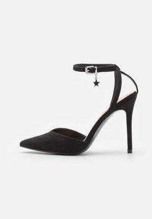 Schwarzer Wildleder-Pumps mit spitzer Schuhspitze, dünnem Knöchelriemen, silberner Schnalle und kleinem sternförmigem Anhänger auf weißem Hintergrund.