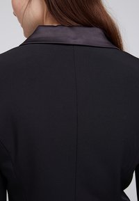 Svart blazer med satinkrage, strukturerad design, slät textur och skräddarsydd passform med en central söm baktill.