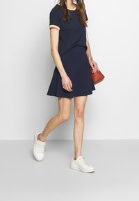 Abito blu navy con maniche corte, caratterizzato da rifiniture rosa e gialle, gonna svasata, abbinato a sneaker bianche e a una piccola borsa a tracolla rossa.