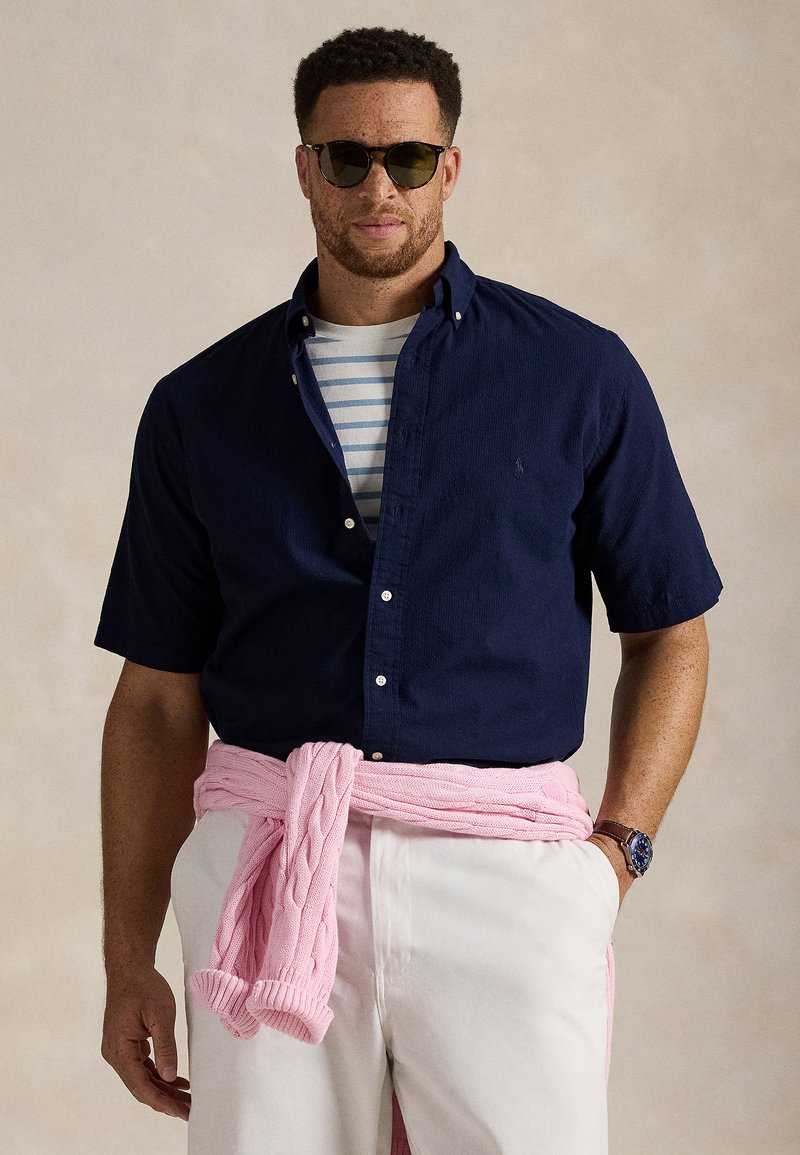 Homme portant des lunettes de soleil, une chemise à manches courtes navy par-dessus un t-shirt rayé, un pantalon blanc et un pull rose noué autour de la taille.