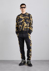 Melnā džemperis ar zelta spirāļu rakstu, savienots ar melnu biksēm ar zelta akcentiem. Valkā melnas un zelta sporta kurpes.