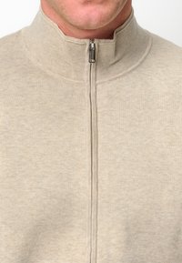 Sudadera de color beige claro con cremallera y cuello alzado, hecha de una tela suave con textura lisa. Cuenta con una cremallera central y un diseño de cuello alto.