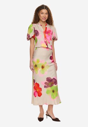 Femme portant une tenue beige deux pièces avec de grands imprimés floraux colorés, des manches bouffantes et des talons slingback noirs, posant devant un fond blanc.