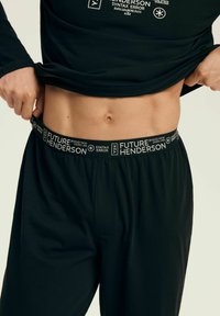 Person lifting black shirt, showing bare midriff and black pants with printed waistband reading "FUTURE HENDERSON" and other text.