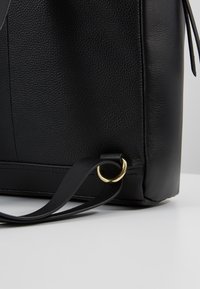 Zwarte leren tas in de hoek met een combinatie van textuur en gladde panelen, gestikte naden, een gouden metalen ring en een bevestigde zwarte schouderband, op een witte ondergrond.