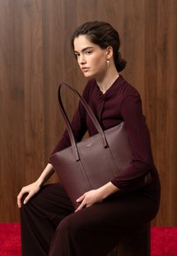 Femme en tenue bordeaux foncé tenant un grand sac fourre-tout en cuir marron, assise contre un mur en bois avec un tapis rouge en arrière-plan.