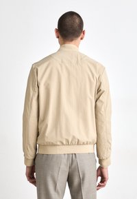 Beige bomberjakke med ribbestrikkede mansjetter og hem, med en glatt tekstur og et minimalistisk design, båret over grå bukser.