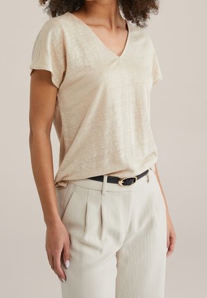 Kobieta ubrana w beżową, krótką bluzkę z dekoltem w serek, wsuniętą w wysokie, kremowe spodnie w kant z czarnym paskiem.