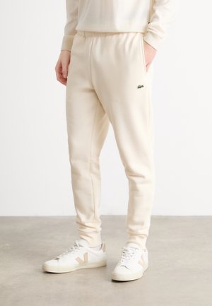 Personne portant un pantalon jogger couleur crème avec un petit logo crocodile et des baskets blanches avec des touches beiges.