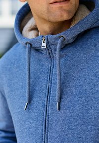 Felpa blu con zip, caratterizzata da un morbido rivestimento interno, cordini regolabili e una zip metallica. Tessuto testurizzato simile al pile.