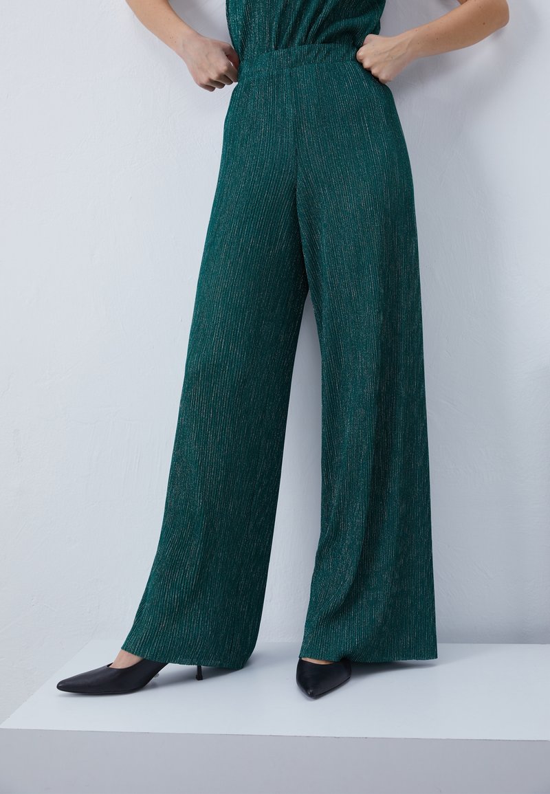 Femme portant un pantalon vert texturé taille haute, jambes larges, et des escarpins noirs à bout pointu, debout sur une plateforme blanche contre un mur blanc.