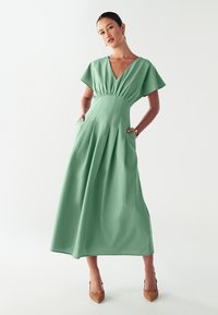 Vestido verde hasta el suelo con mangas cortas y onduladas, escote en V, cintura ajustada y bolsillos laterales; hecho de una tela suave con un diseño plisado.