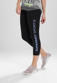 Černé legíny s světle modrým potiskem "SUPERDRY SPORT" podél levé nohy, nošené se světle růžovými sportovními botami. Materiál vypadá elasticky.