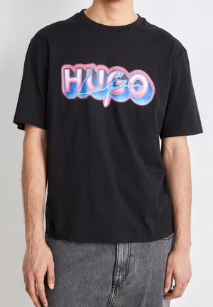 Mann trägt schwarzes T-Shirt mit großem buntem „HUGO“-Schriftzug in Pink und Blau, kombiniert mit grauen Jeans aus Denim.