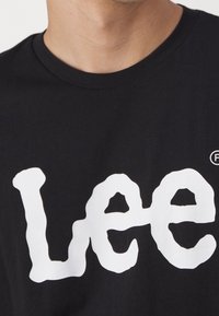 Gros plan sur une personne portant un t-shirt noir avec un grand texte blanc déformé épelant "Lee" sur la poitrine.