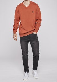 V-ringad, långärmad tröja i bränd orange, tillverkad av stickat material. Matcher med mörka jeans och vita sneakers. Broderad logotypdetalj på bröstet.