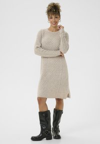Glimlachende vrouw met krullend haar, gekleed in een beige gebreide jurk en zwarte laarzen tot de knie van leer, poserend tegen een witte achtergrond.