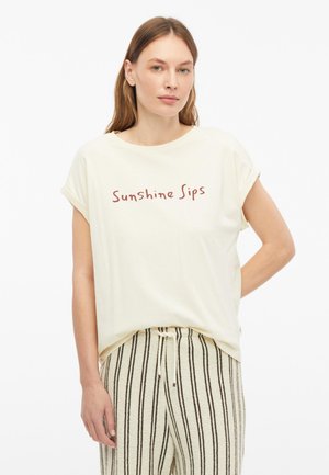 Eine Frau trägt ein cremefarbenes T-Shirt mit lockerer Passform und hochgekrempelten Ärmeln mit dem Schriftzug "Sunshine Sips", kombiniert mit beige-schwarz gestreiften Hosen.
