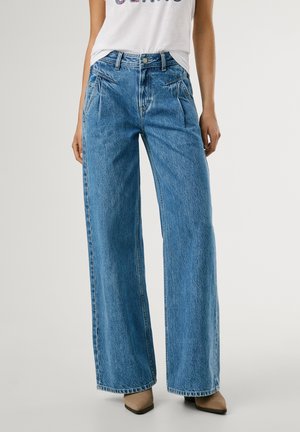 Brede blauwe denimbroeken met een hoge taille, voorzien van voorzakken en een vervaagde textuur. Gestyleerd met een wit graphic T-shirt en hakken.