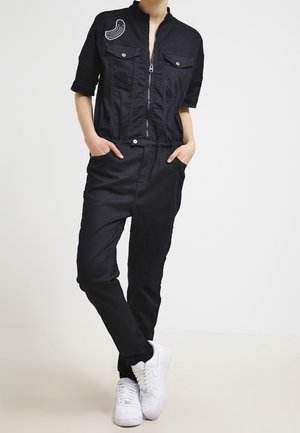 Person iført en sort kortærmet jumpsuit med lynlås og brystlommer samt hvide sneakers, stående med hænderne i lommerne.