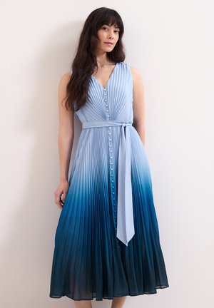 Vrouw met lang donker haar, gekleed in een mouwloze, geplooide blauwe ombre jurk met knopen en een gestrikte tailleriem, staand tegen een effen lichte muur.