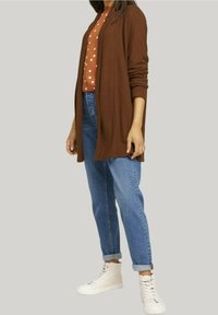 Brun lång kofta lager på lager över en prickig brun topp, kombinerad med blå jeans och vita högtoppsneakers. Enkelt design, avslappnad passform.