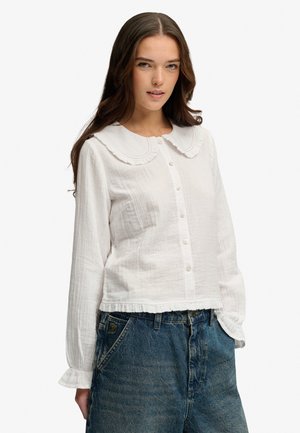 Kvinde med langt brunt hår iført en hvid knapbluset med en bred flæset krave og blå højttaljede jeans.