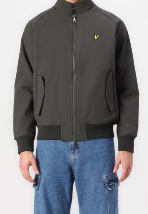 Homme portant une veste zippée gris foncé avec col montant et logo oiseau jaune, associée à un jean cargo bleu, debout devant un fond blanc.