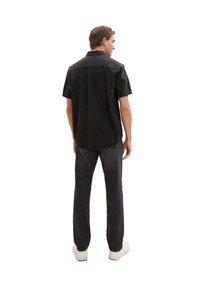 Camicia in denim nera a maniche corte con una piega sul retro, indossata con jeans scuri e sneakers bianche, che mette in risalto un look casual e un design semplice.