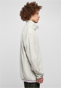 Vaaleanharmaa fleece-pullover, jossa on korkea kaulus, puolivälin vetoketju ja erottuvat kankaat hihoissa. Pehmeä rakenne ja rento istuvuus.