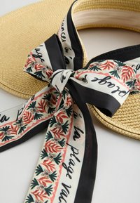 Visière en paille à large bord nouée avec un foulard à motif floral noir, blanc et rouge comportant des accents de texte en écriture cursive.