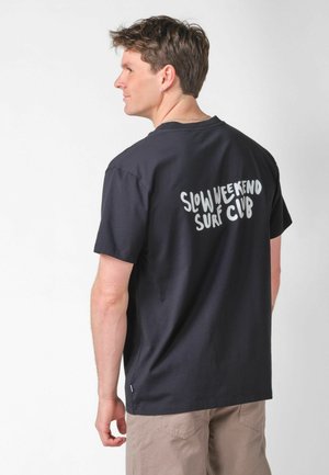 Junger Mann in schwarzem T-Shirt mit dem Schriftzug "SLOW WEEKEND SURF CLUB" auf dem Rücken, trägt beige Shorts und steht vor einem einfarbigen weißen Hintergrund.