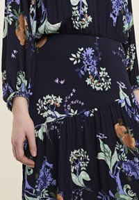 Blumenmuster-Kleid aus dunkelblauem Stoff; verfügt über geraffte Ärmel und einen fließenden Rock mit hellgrünen, lavendelfarbenen und braunen botanischen Details.