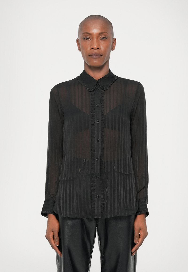 Schwarze transparente Bluse mit vertikalen Streifen, geknöpftem Vorderteil, Rüschenkragen und -manschetten. Kombiniert mit schwarzen Lederhosen.