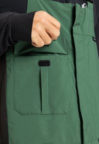 Hand met een zwarte mouw die de klittenbandsluiting van een grote groene zak op een groen en zwart kledingstuk vastmaakt.