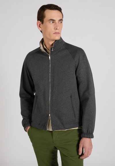 Uomo con giacca grigia scura con cerniera e pantaloni verde oliva, guarda di lato con le mani parzialmente nelle tasche su sfondo neutro.