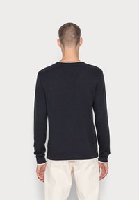 Mężczyzna stojący tyłem, ubrany w czarny sweter z długim rękawem i kremowe spodnie, na tle jasnego, jednolitego tła.