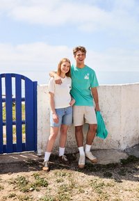 Pareja al aire libre junto a una puerta azul; mujer con una camiseta crema y shorts de mezclilla, hombre con una camisa turquesa y shorts beiges, ambos con zapatillas.