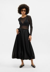 Fekete hálós, hosszú ujjú felső, amelyet egy tágas fekete selyem szoknyával párosítanak. Kiegészítők között gömbölyű napszemüveg és díszített lapos cipők találhatóak.