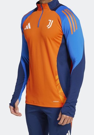 Homme portant un haut de sport à manches longues avec des panneaux orange et bleu, pantalon bleu, et des logos sur la poitrine et les manches, debout, mains détendues.