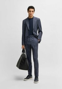 Dark blue, textured suit with a tailored fit, paired with a navy shirt and black sneakers. Holding a black bag with dual handles.