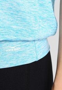 Top deportivo azul claro con un acabado texturizado, presenta un dobladillo redondeado y el logo "VENICE BEACH" en blanco en la parte inferior.